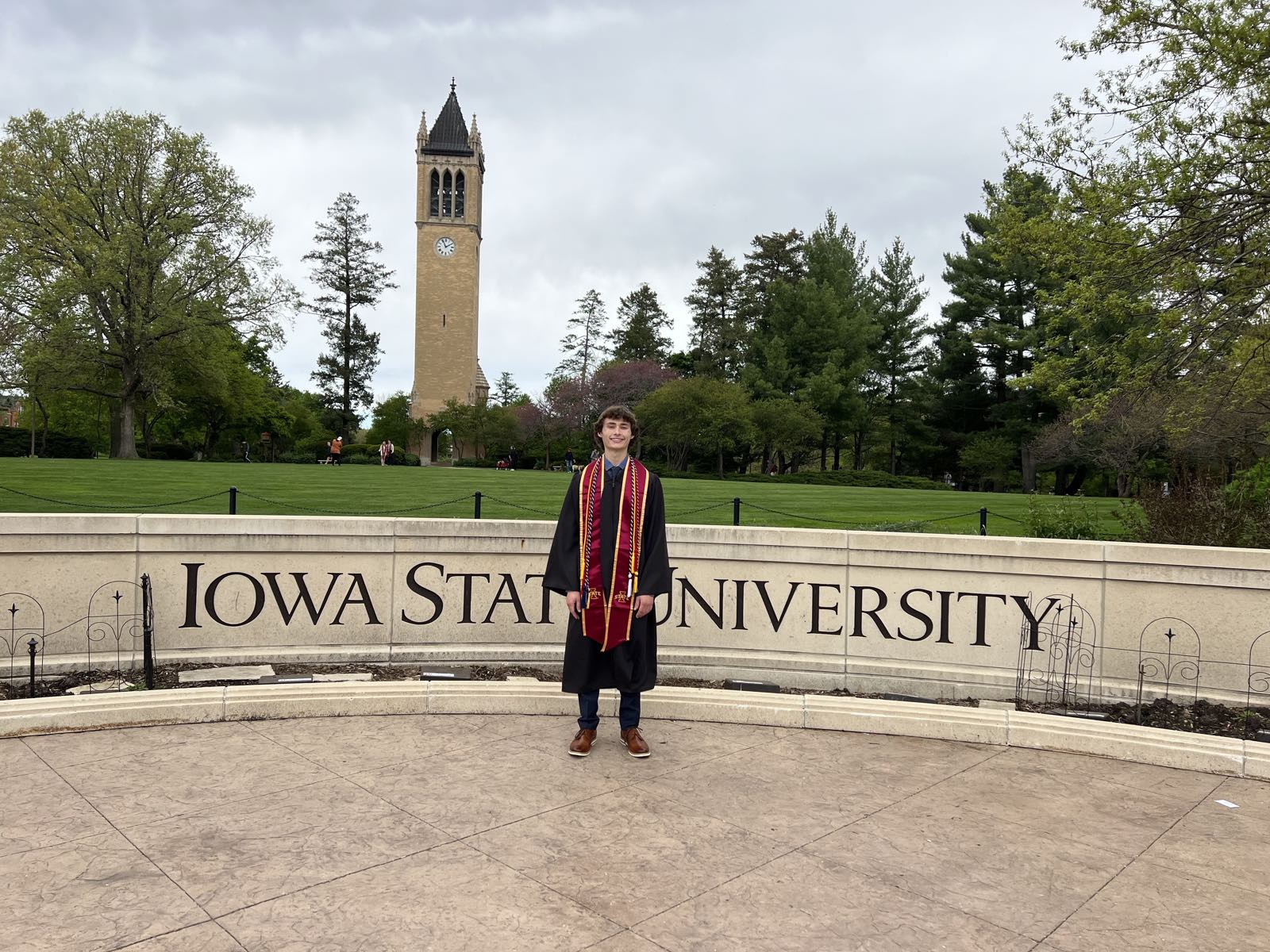 Michael in graduation robes in front of the Iowa State University sign, campanile behind